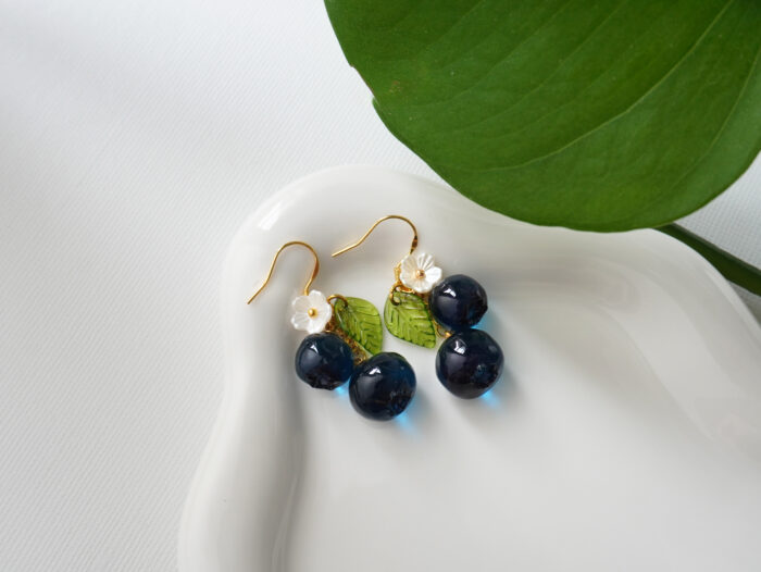 Close-up of handmade blueberry resin dangle earrings with green enamel leaf and white flower