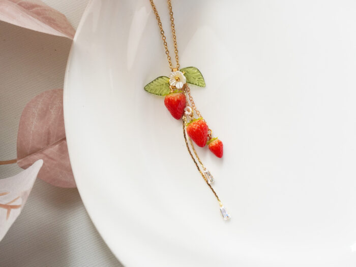 Macro view of three glossy polymer clay strawberries and gold chain with faceted crystal ends on a white plate