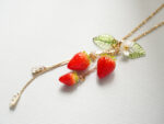 Flat lay of a strawberry pendant necklace showing leaf, flower, and crystal charms spread on a textured white surface
