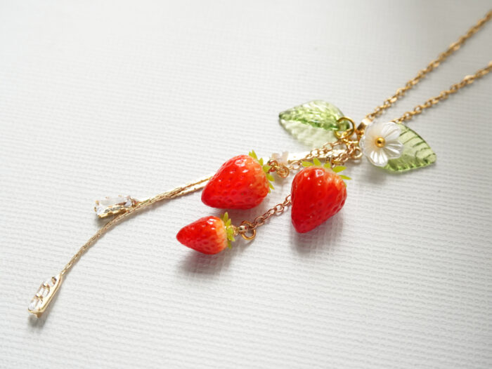 Flat lay of a strawberry pendant necklace showing leaf, flower, and crystal charms spread on a textured white surface