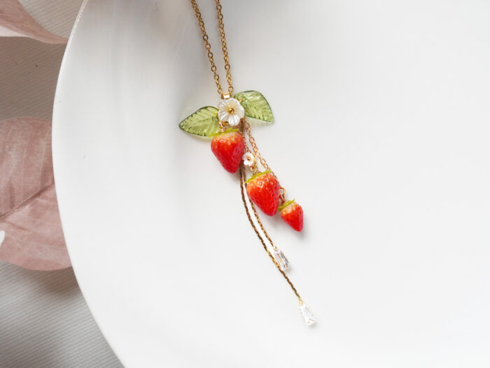 Polymer clay strawberry necklace laid on a white plate showing the full pendant, gold chain, and dangling crystal strands