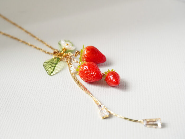 Angled macro shot of polymer clay strawberry charms with a green leaf and crystal drop chain on a textured surface
