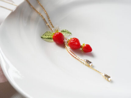 Close-up diagonal view of three polymer clay strawberries with a flower charm and two zircon crystal drops on a white plate