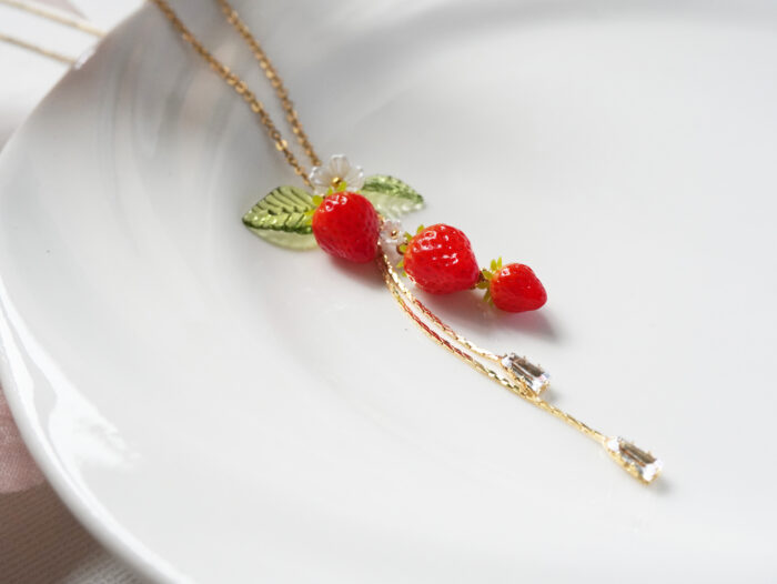 Close-up diagonal view of three polymer clay strawberries with a flower charm and two zircon crystal drops on a white plate