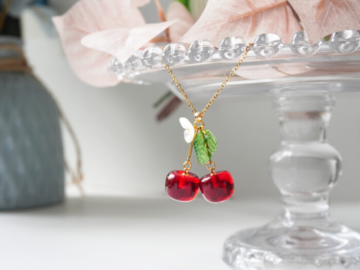 Close-up of a resin cherry pendant necklace draped over a glass cake stand edge with dried pink leaves
