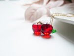 Detail view of two glossy red resin cherry charms and green leaf beside a white ceramic dish